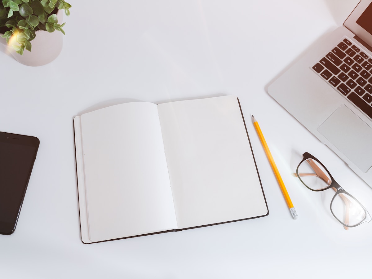 Notebook, pencil, phone, and laptop arranged on a white desk.
